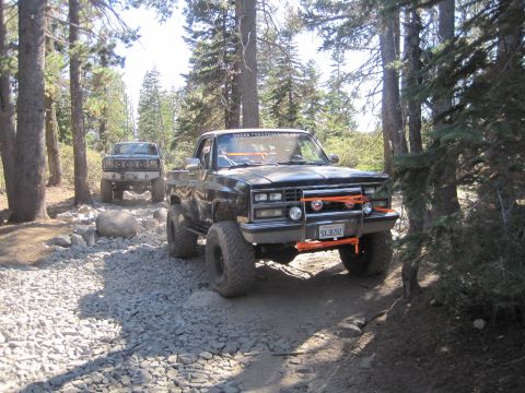 1989 Ford Bronco - Two off-road pickup trucks navigate a rocky trail through a forest.