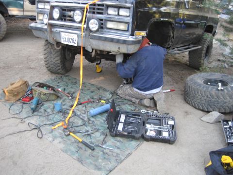 1989 Ford Bronco - A person works on the underside of a black pickup truck, with tools laid out on a camouflage tarp.
