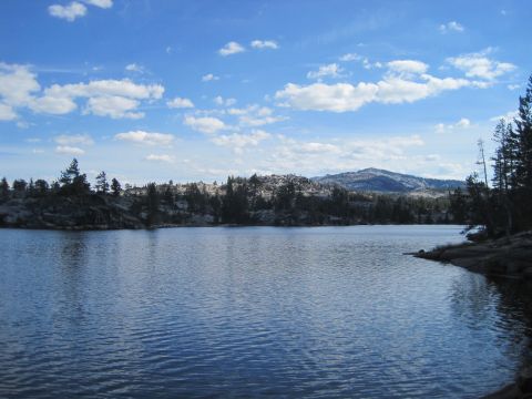 1989 Ford Bronco - A calm lake reflects the blue sky and scattered clouds, with a backdrop of trees and mountains.