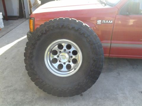 1968 Ford Bronco - A large tire and chrome rim are positioned in front of a red truck with 'RAM' badging.