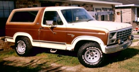 1980 Ford Bronco - Front 3/4 side view of a brown and white two-tone full-size Ford Bronco parked on grass in front of a brick house, wearing a white removabl…