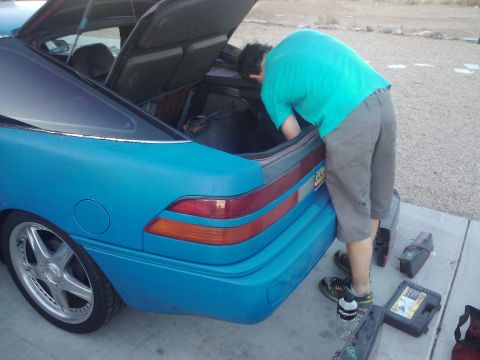 1995 Ford Probe - A person is searching through the trunk of a blue coupe, with various tools laid out on the ground nearby.