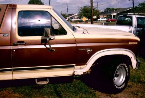 1980 Ford Bronco - Passenger-side profile of a two-tone brown and cream Ford Bronco parked on grass by a residential street, showing the 'BRONCO' fender emble…