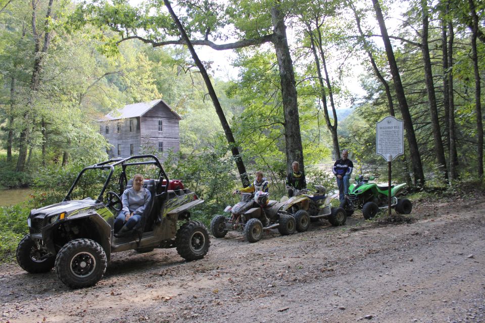 Four ATVs parked on a dirt road near an old wooden building and trees. People are on or near the ATVs