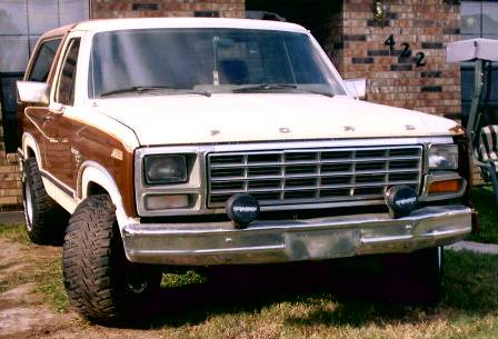 1980 Ford Bronco - Front 3/4 view of a brown and white two-tone full-size Ford Bronco parked on grass beside a brick house numbered 422, showing the FORD bloc…