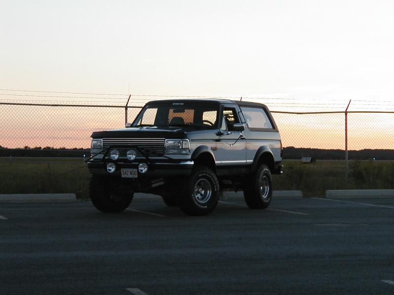 Lifted two-tone Ford Bronco with bumper-mounted driving lights parked in a lot at sunset