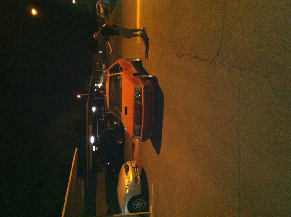 Orange sports car parked at night, with a person standing nearby. Another car is visible in front