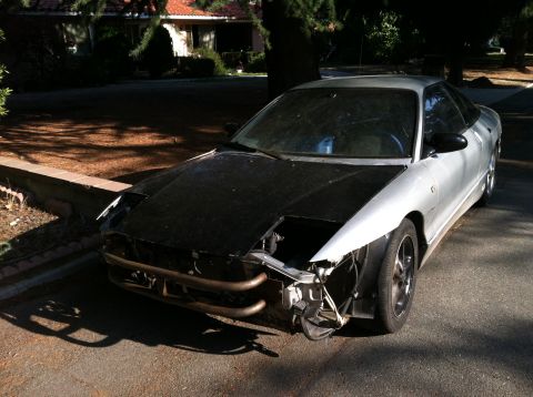 1993 Ford Probe - A silver 1993 Ford Probe with a black hood and a damaged front end sits parked on a residential street.
