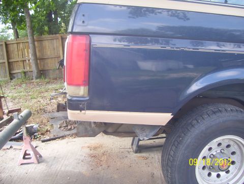 1990 Ford Bronco - Rear quarter of a blue two-tone SUV with amber-and-red taillight and dual rear wheels raised on jack stands.