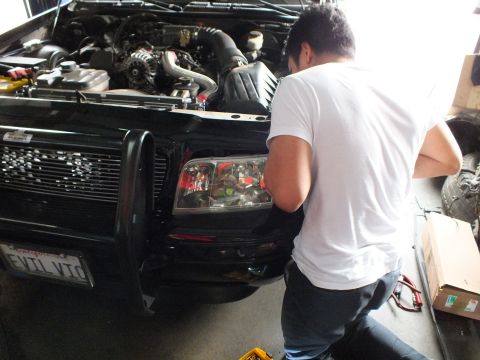 2003 Ford Crown Victoria - Mechanic leans into the headlight of a black grille-guard sedan plated EVIL VIC inside a shop.