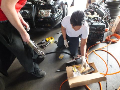 2003 Ford Crown Victoria - Two mechanics use pneumatic tools on the floor near a black sedan missing its headlights.