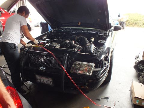 2003 Ford Crown Victoria - Mechanic works in the engine bay of a black brush-guard sedan plated EVIL VTC, red jumper attached.