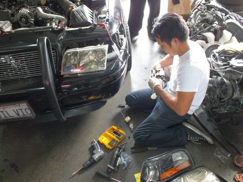 2003 Ford Crown Victoria - Technician seated on garage floor assembling a headlight while a black push-bar sedan sits in the background