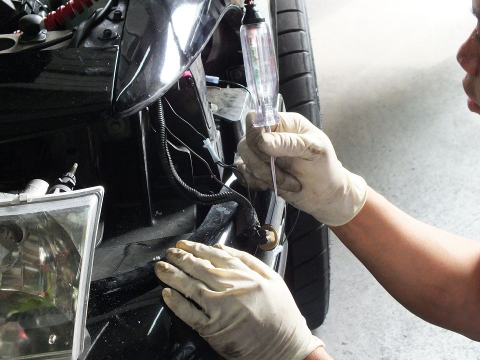 Gloved hands using a circuit test light probe on wiring near a black car front bumper and headlight