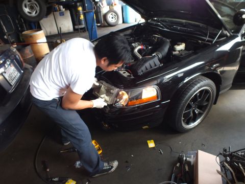2003 Ford Crown Victoria - Technician works at the front fender of a black sedan with open hood, cold-air intake and dark alloy wheels visible