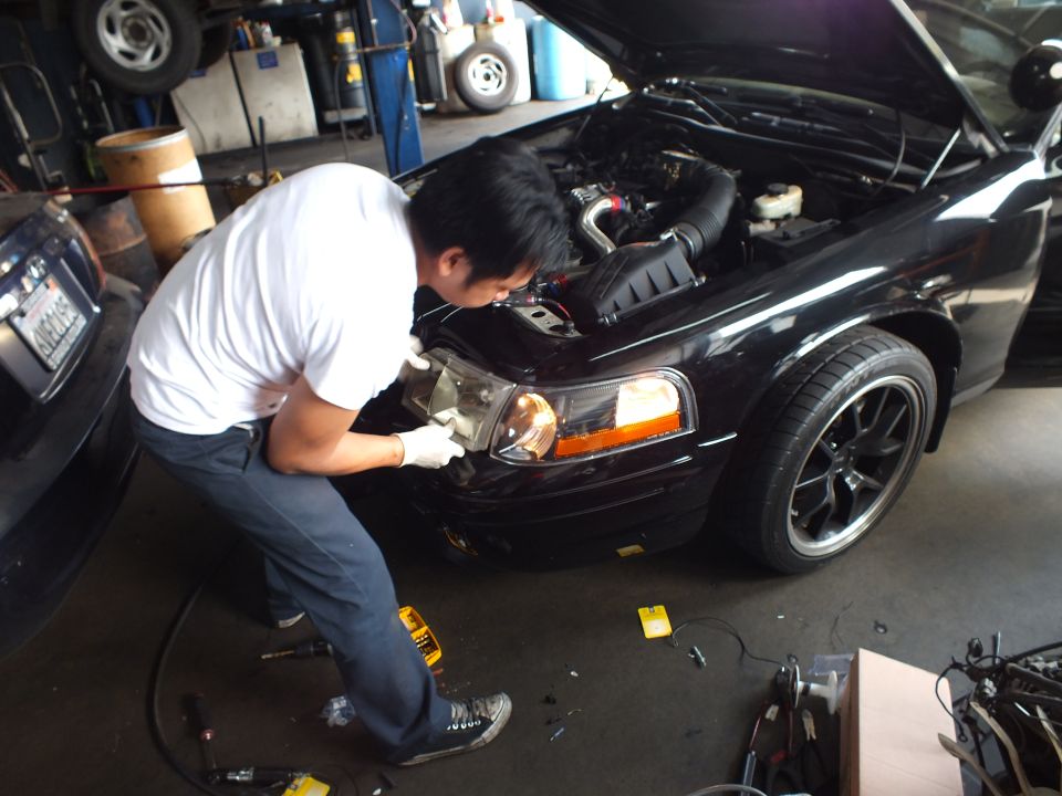 Man in white shirt bending over front fender of black sedan with hood up and black aftermarket wheels in shop