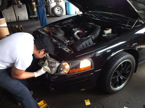 2003 Ford Crown Victoria - Worker kneels at the front of a dark sedan, one headlight illuminated, engine bay open with intake tubing