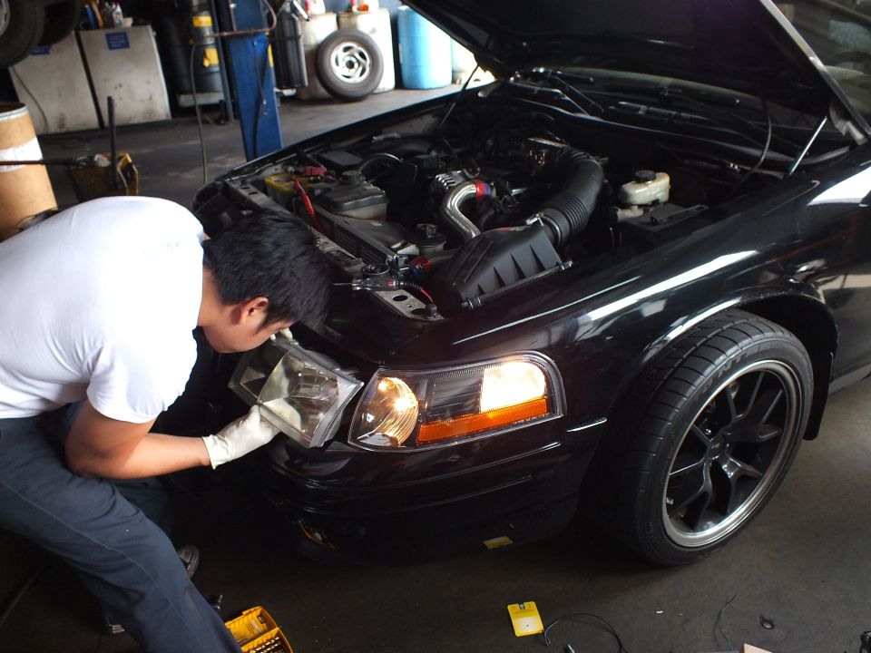 Man crouched at front corner of dark sedan with hood open, lit headlight and amber turn signal, black wheels