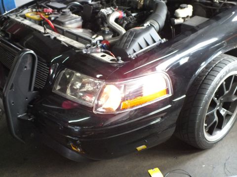 2003 Ford Crown Victoria - Open engine bay of a dark sedan showing front-mount intercooler and intake piping with black multi-spoke wheel