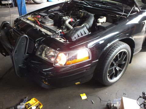 2003 Ford Crown Victoria - Engine compartment of a dark sedan exposed with hood up, headlight on, tools scattered on the garage floor