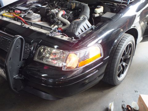 2003 Ford Crown Victoria - Front corner of a black Ford Crown Victoria in a garage, hood raised to show the engine; one headlight and amber turn signal are lit.