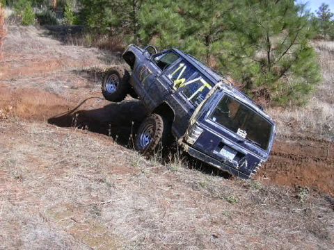 1988 Ford Bronco - This dark blue SUV, equipped with a yellow roll cage, navigates a challenging off-road trail, with one wheel lifted off the ground.