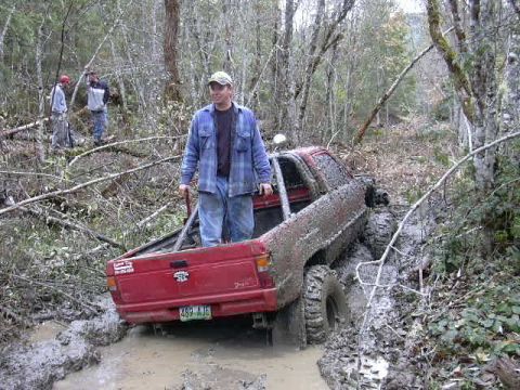 1988 Ford Bronco - A red pickup truck is deeply stuck in mud on a wooded trail, with a man standing in the bed.