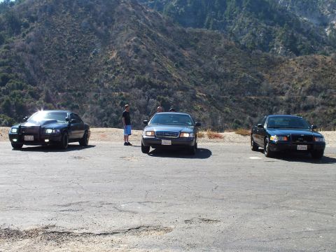 2003 Ford Crown Victoria - Trio of dark sedans lined up at a hillside turnout, one person standing center, dry hills in background