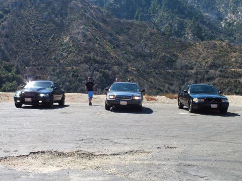 2003 Ford Crown Victoria - Three vehicles parked across a paved turnout, headlights lit, one person crossing, forested hills behind