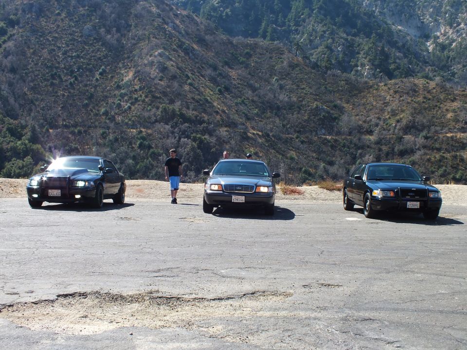 Three dark sedans parked on cracked asphalt lot with headlights on, person walking between, green hillside behind
