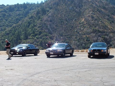 2003 Ford Crown Victoria - Three cars lined across cracked pavement with sun flaring off front ends, people standing by, green ridge behind