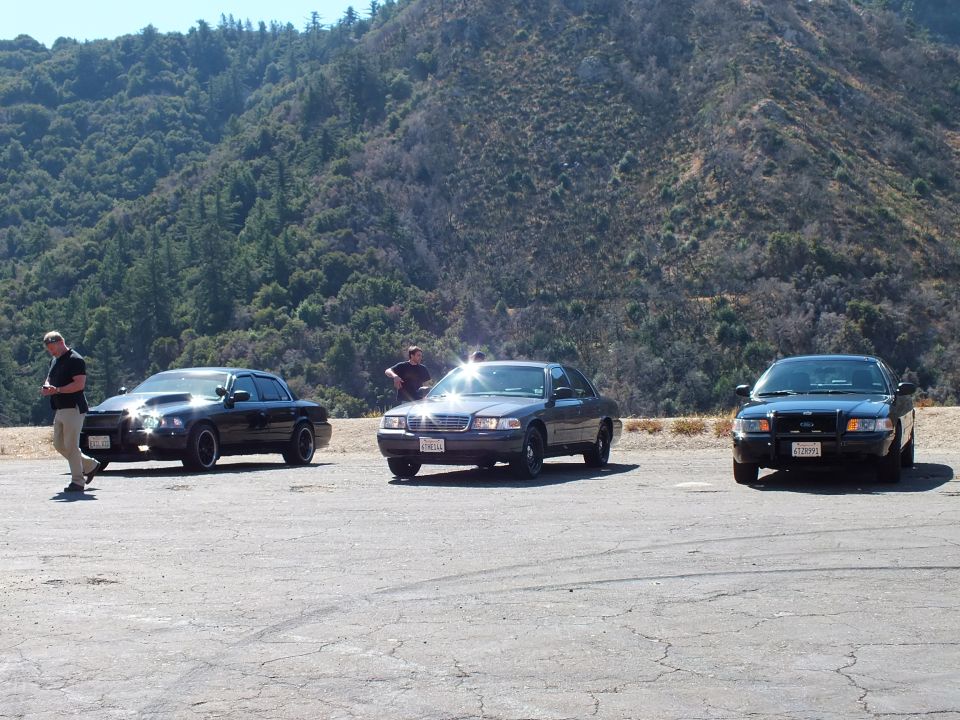 Three dark sedans on paved lot with sun glare on grilles, two men standing nearby, wooded mountain behind