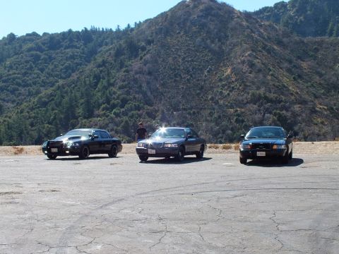 2003 Ford Crown Victoria - Trio of dark cars on a paved lookout, bright sun reflection on the middle vehicle, green hills behind