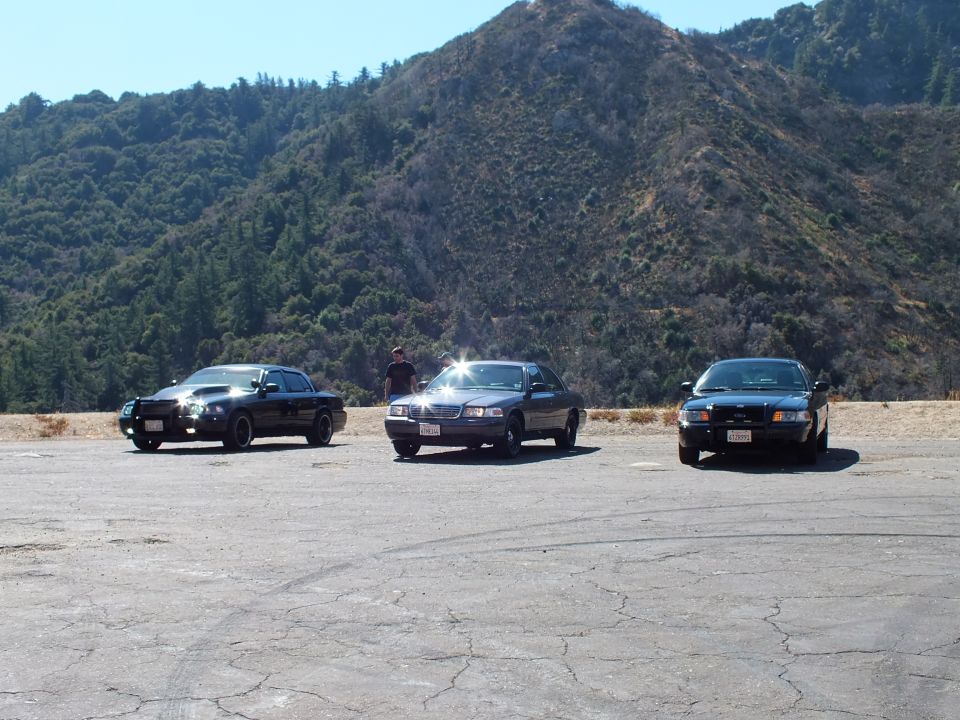 Three dark sedans parked across a gravel lot, sun glare on center car, forested mountainside under blue sky