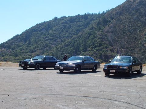 2003 Ford Crown Victoria - Three full-size sedans across a paved turnout, a silver one centered between two black cars, wooded ridge behind