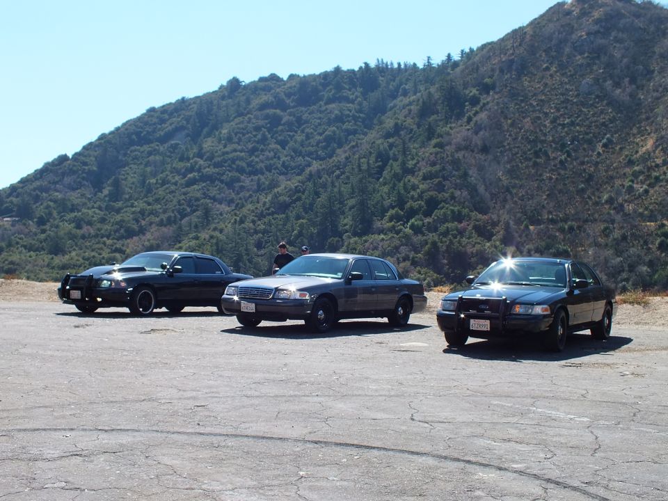 Three dark sedans lined across gravel lot, silver Ford center, two black sedans flanking, green mountainside behind