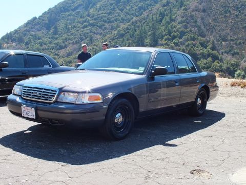 2003 Ford Crown Victoria - Slate-gray Crown Victoria sedan sits on a gravel turnout below a brushy ridge, two men standing behind it.