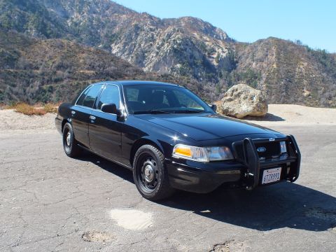 2003 Ford Crown Victoria - Black Crown Victoria Police Interceptor with front push bar parked on pavement before rocky chaparral hills.