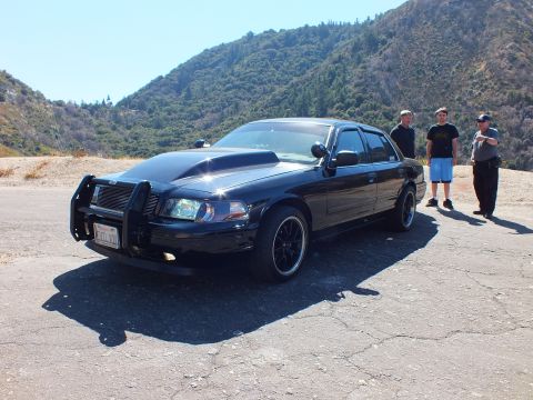 2003 Ford Crown Victoria - Customized black Crown Victoria with cowl hood and push bar on a turnout while three men stand nearby.