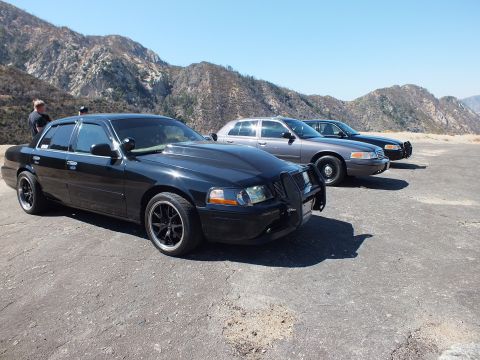 2003 Ford Crown Victoria - Lowered black Mercury Grand Marquis on dark wheels leads two parked Ford sedans on a gravel pullout.