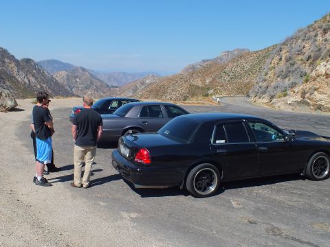 2003 Ford Crown Victoria - Two men talk beside parked sedans, a black Crown Victoria in front, on a turnout overlooking ranges.