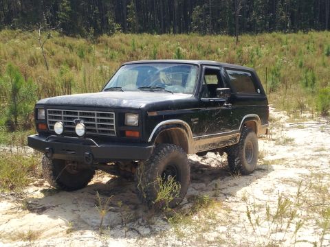 1986 Ford Bronco - 1986 Ford Bronco parked off-road in a wooded area with sandy ground.