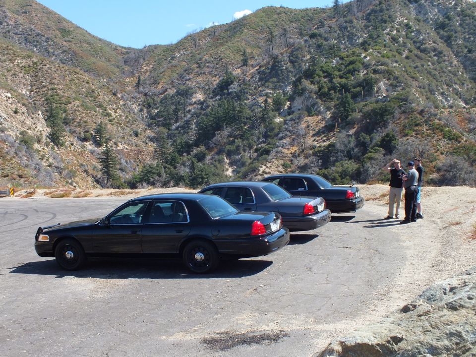 Four black Ford sedans parked in row on mountain pullout with men at right