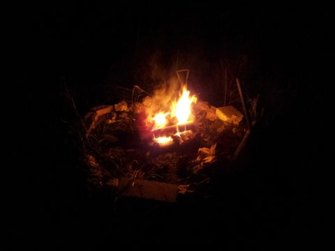 1986 Ford Bronco - A campfire glows brightly in the darkness, illuminating the surrounding rocks and sparse vegetation.