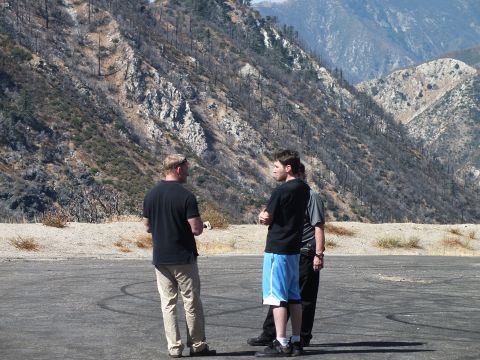 2003 Ford Crown Victoria - group converses on an open paved pad with burned brush and rocky ridges behind them