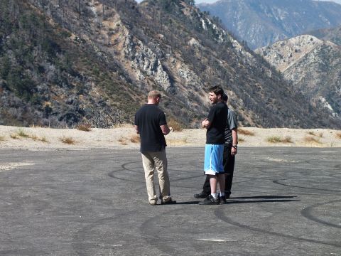 2003 Ford Crown Victoria - Men gather on a wide paved turnout scarred with tire marks below burned hillsides