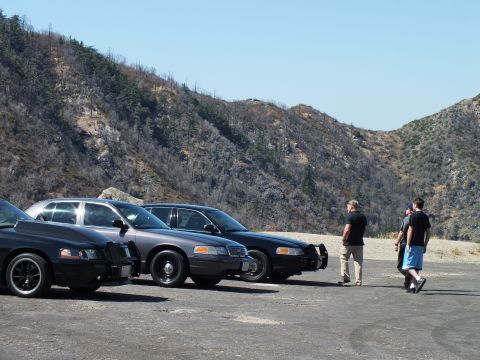 2003 Ford Crown Victoria - Two men stroll past a row of dark sedans on an open gravel pad beneath brushy hills