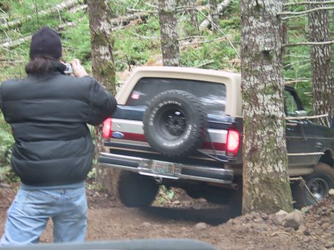 1991 Ford Bronco - A person photographs a 1991 Ford Bronco navigating a wooded trail, with another vehicle visible behind.