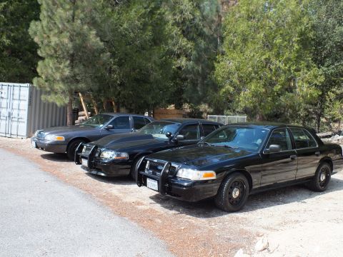 2003 Ford Crown Victoria - gray sedan and two black sedans with push bars line a gravel drive by a shipping container