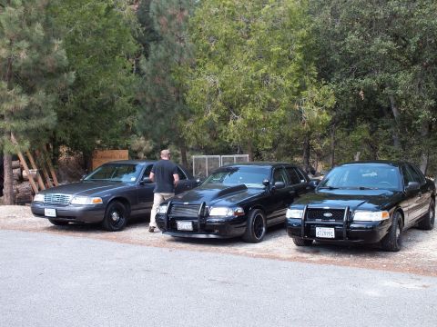 2003 Ford Crown Victoria - man stands at a sedan door beside two black push-bar cars on a gravel forest drive